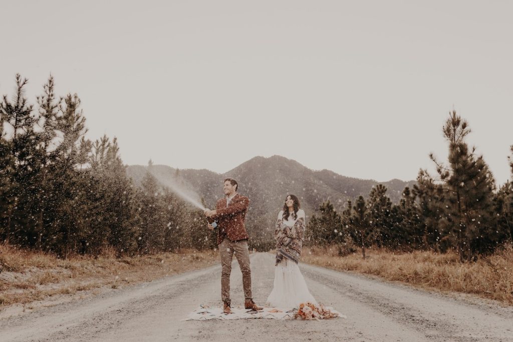 Bride and groom celebrating with champagne spray on country road with mountain in the background